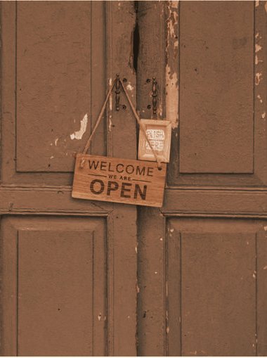 A wooden "Welcome, we are open" sign hanging on a weathered teal door